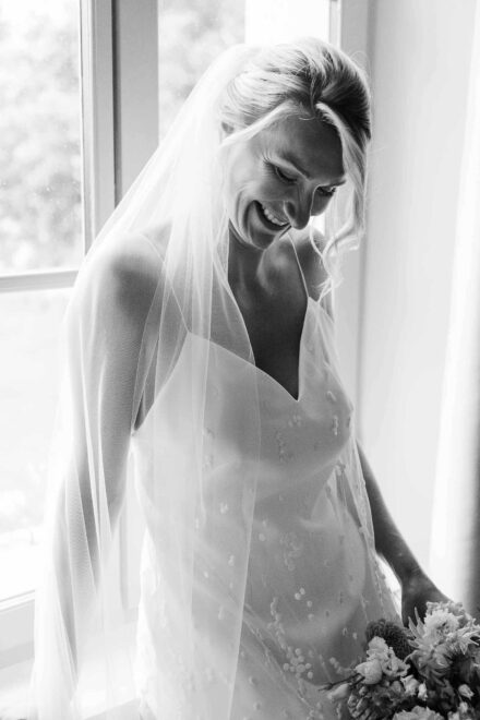 La novia, con velo y vestido de tirantes bordado, sonríe mirando hacia abajo junto a la ventana, en blanco y negro