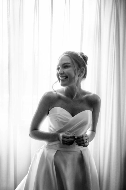 La novia, con vestido palabra de honor, sonríe frente a unas cortinas a contraluz, en blanco y negro