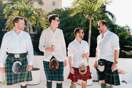 Cuatro amigos con kilt y camisa blanca se ríen con latas de cerveza en una terraza entre palmeras