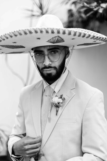 El novio, con sombrero de charro y gafas redondas, se abrocha la chaqueta mirando a cámara, en blanco y negro