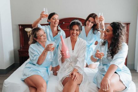 La novia, con una botella rosa en la mano, ríe en la cama entre cuatro mujeres con batas celestes que brindan