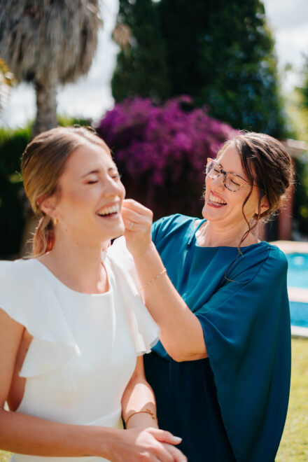 La novia se ríe mientras una invitada con gafas y vestido azul petróleo le retoca el pelo en el jardín