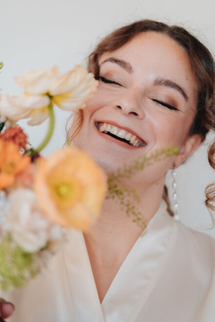 La novia, en bata y con los ojos cerrados, se ríe detrás de un ramo de amapolas desenfocado