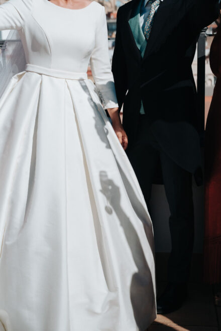 La sombra de una mano con una copa cae sobre la falda de satén de la novia, al lado del novio de chaqué, en color