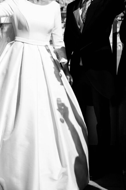 La sombra de una mano con una copa se proyecta sobre el vestido de la novia, junto al novio de chaqué, en blanco y negro