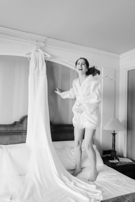 La novia, en bata, salta riendo sobre la cama junto al vestido de novia colgado, en blanco y negro