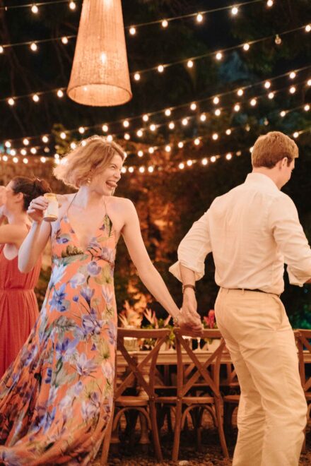 Una invitada con vestido largo de flores baila riendo de la mano de un chico de camisa blanca bajo guirnaldas de luces