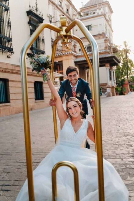 La novia levanta el ramo en un carro portaequipajes dorado que empuja el novio, frente al Hotel Alfonso XIII (Sevilla)