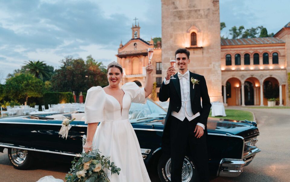 Bride and groom toasting with champagne beside a vintage car at Castillo de la Monclova in Seville