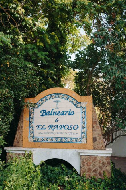 Cartel de azulejos a la entrada del Balneario El Raposo, rodeado de árboles y enredaderas