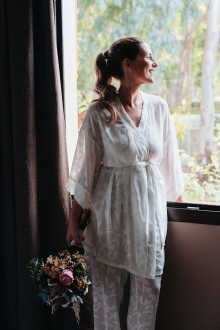 La novia, en pijama blanco, sonríe junto a la ventana con el ramo en la mano durante los preparativos