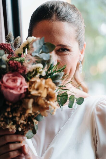 La novia se tapa media cara con el ramo de flores secas y mira a cámara con un solo ojo
