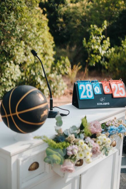 Balón negro con líneas doradas, micrófono y marcador de tanteo sobre un mueble blanco con flores