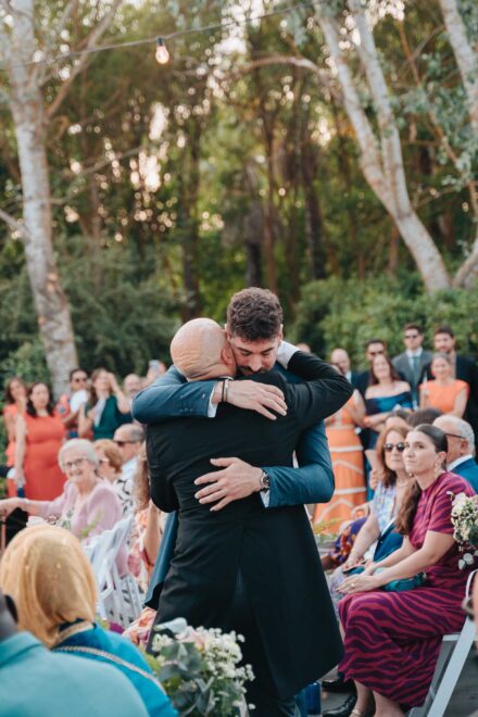 El novio abraza a un hombre de chaqué en el pasillo de la ceremonia, ante los invitados sentados