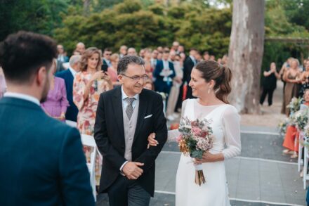 La novia avanza por el pasillo del brazo de un hombre con chaqué y se sonríen, con el novio de espaldas