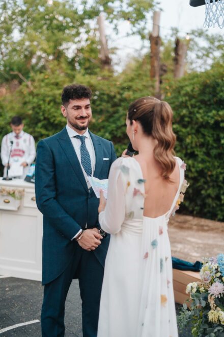 El novio sonríe mientras la novia, de espaldas, le lee sus votos con el micrófono en la mano