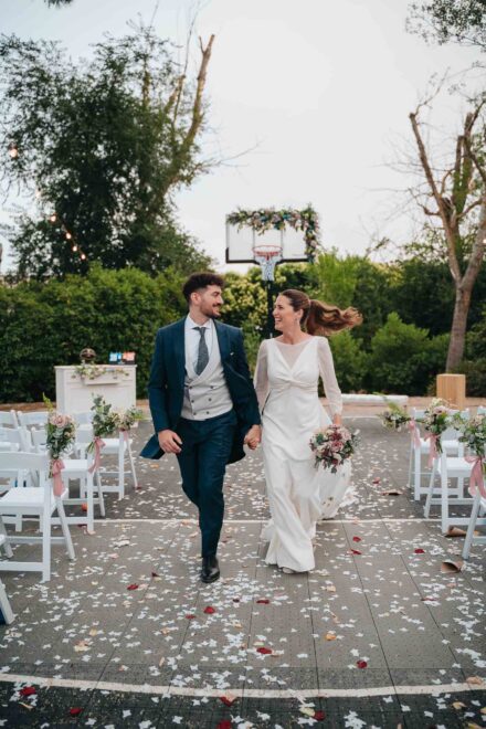 Los novios caminan de la mano por el pasillo de sillas blancas, ella con la coleta al viento