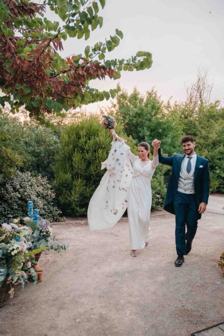 La novia alza el ramo y la cola del vestido mientras camina con el novio, las manos unidas en alto