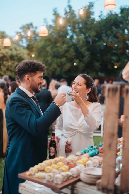 Los novios se ríen, cada uno con una pieza de sushi en la mano, junto a la mesa del cóctel bajo lámparas de mimbre