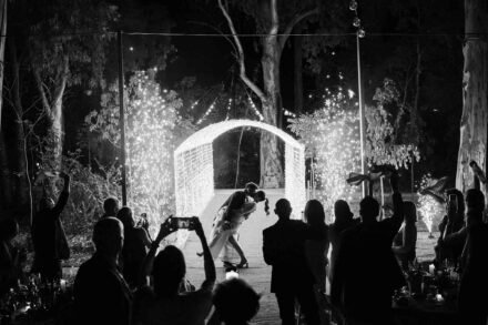 En blanco y negro, el novio inclina a la novia para besarla bajo un arco de luces, entre chispas e invitados
