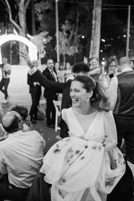 En blanco y negro, la novia ríe recogiéndose la falda entre los invitados mientras el novio saluda con el brazo en alto