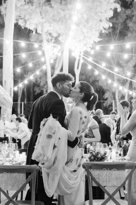 En blanco y negro, los novios se besan de pie entre las mesas del banquete, bajo guirnaldas de bombillas