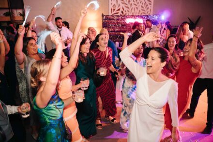 La novia baila con el brazo en alto rodeada de invitadas que cantan y ríen en la fiesta, bajo luces rosas