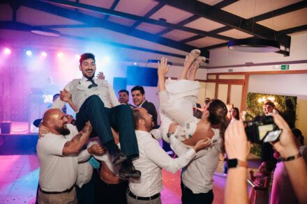 Los invitados levantan en volandas a la vez al novio y a la novia en la fiesta, mientras alguien los enfoca con una cámara
