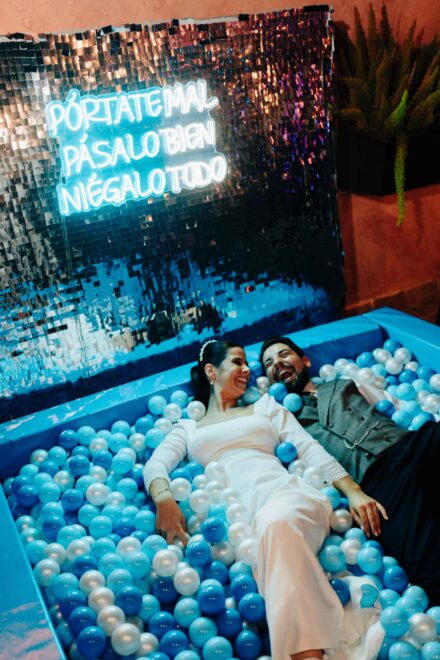 Los novios ríen tumbados en una piscina de bolas bajo el neón «Pórtate mal, pásalo bien, niégalo todo»