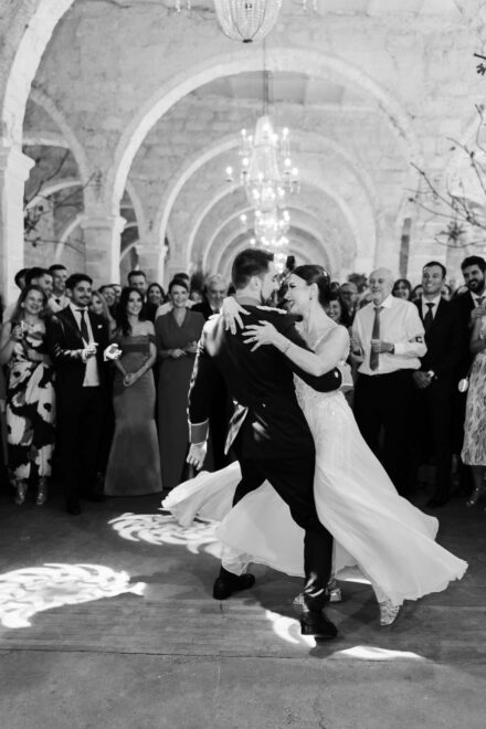 El novio, de uniforme, baila con la novia bajo arcos de piedra, rodeados de invitados, en blanco y negro