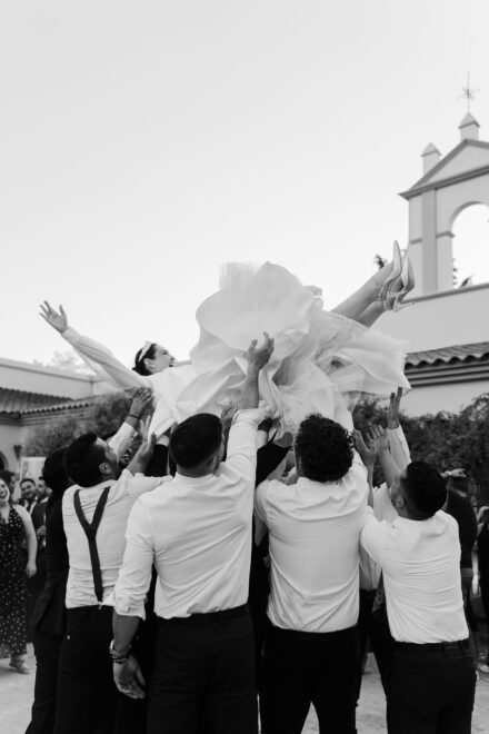 Un grupo de hombres en camisa blanca lanza a la novia por los aires frente a una espadaña, en blanco y negro