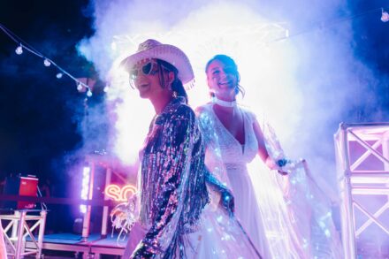 La novia, con capa iridiscente, ríe entre humo violeta junto a una mujer de blanco con sombrero vaquero