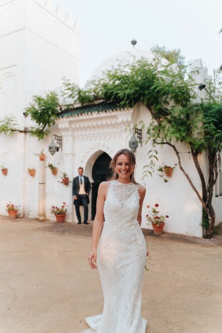La novia sonríe en primer plano mientras el novio espera junto a una puerta en arco de una fachada blanca