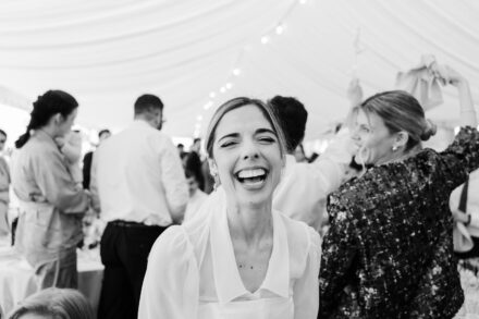 La novia se ríe a carcajadas con los ojos entornados mientras los invitados bailan bajo la carpa, en blanco y negro