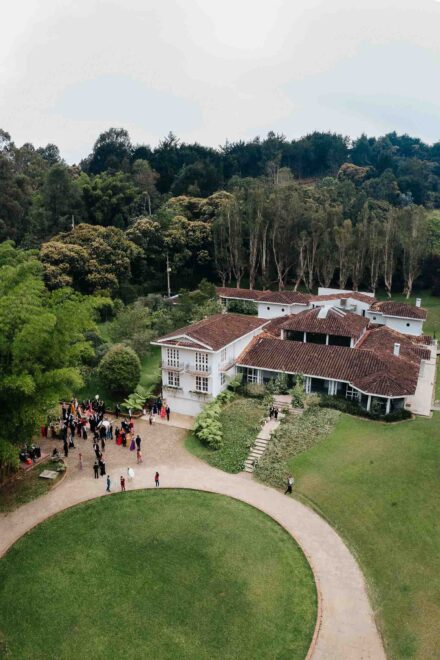 Vista aérea de una casa blanca de tejados rojos entre el bosque, con los invitados reunidos junto a un césped circular
