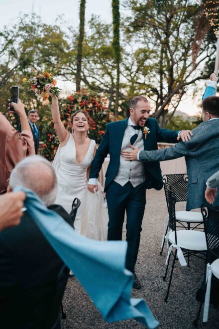 Los novios entran riendo al banquete, ella con el ramo en alto, mientras un invitado agita una servilleta azul