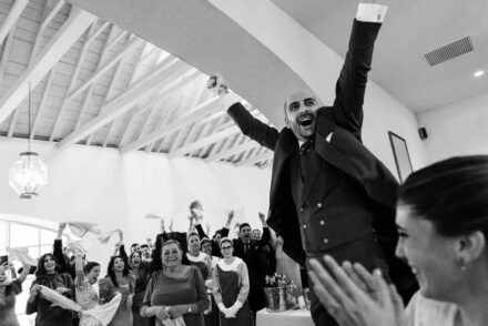 Un invitado salta con los brazos en alto mientras los demás agitan servilletas en el salón, en blanco y negro